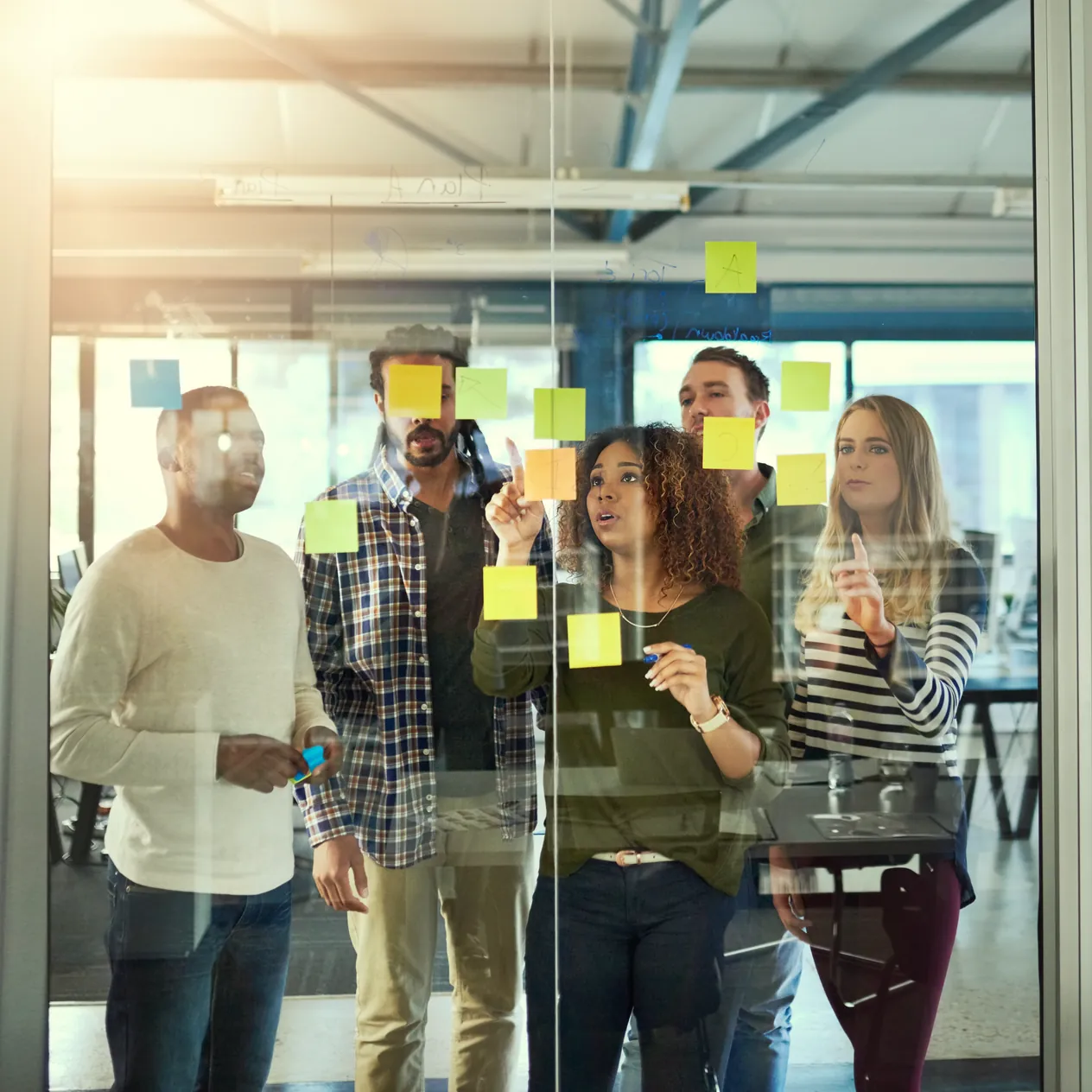 Five colleagues seen through a glass wall covered in sticky notes, adding another to it from the far side