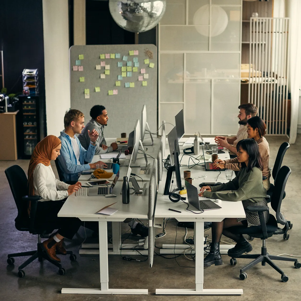 Programmers at a long shared desk, each at their own monitor, a wall of sticky notes behind them