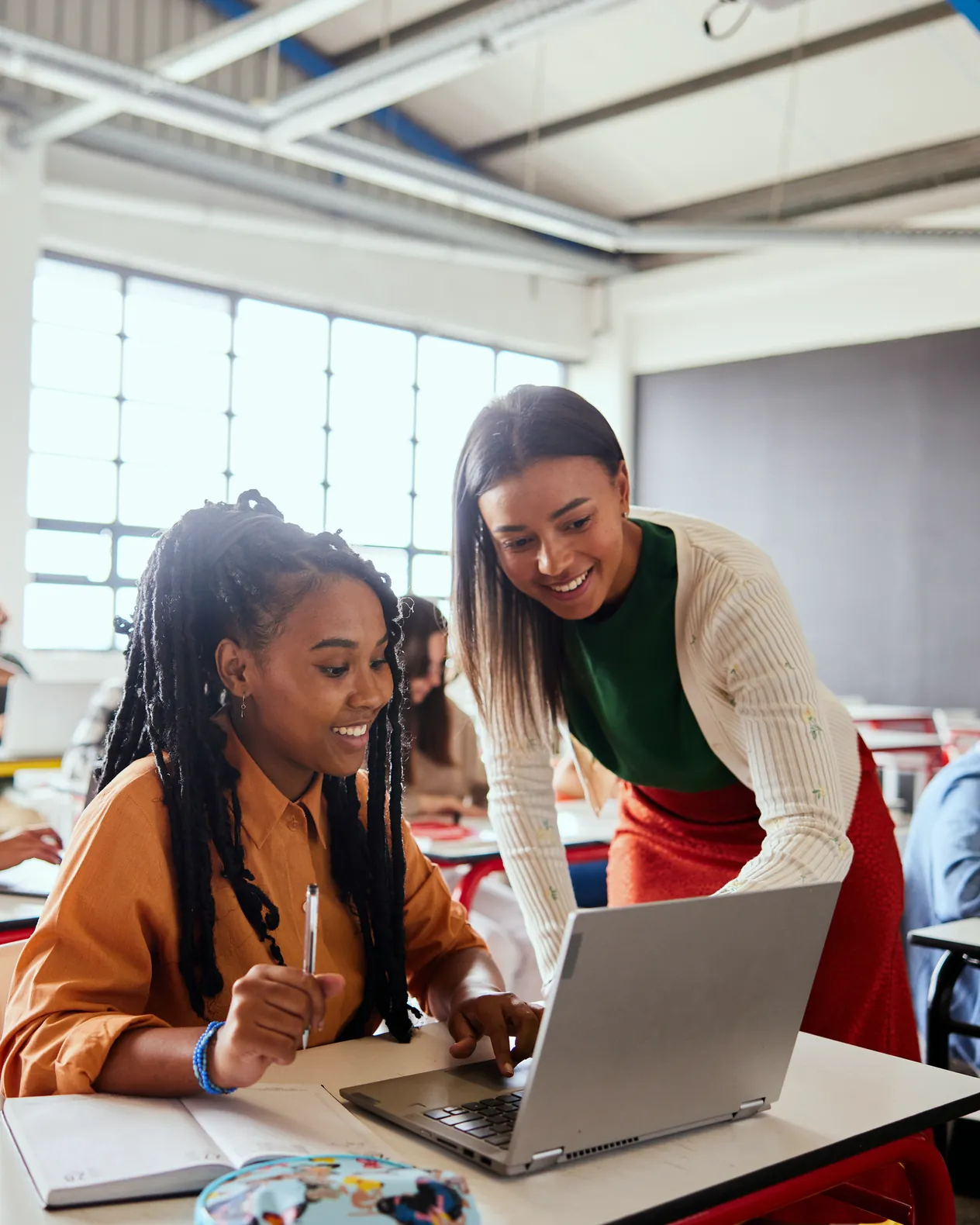 A teacher leaning over a student at a laptop, both smiling at the screen